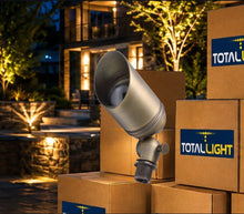 Load image into Gallery viewer, A brass metal outdoor landscape light fixture shown leaning against stacked cardboard boxes with visible TOTALLIGHT branding, set in front of a warmly lit house at night.