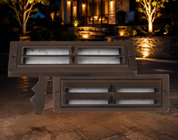 Two rectangular outdoor LED landscape light fixtures in a dark bronze finish, shown against a nighttime patio backdrop with a house and trees in the background.