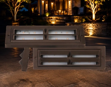 Two rectangular outdoor LED landscape light fixtures in a dark bronze finish, shown against a nighttime patio backdrop with a house and trees in the background.